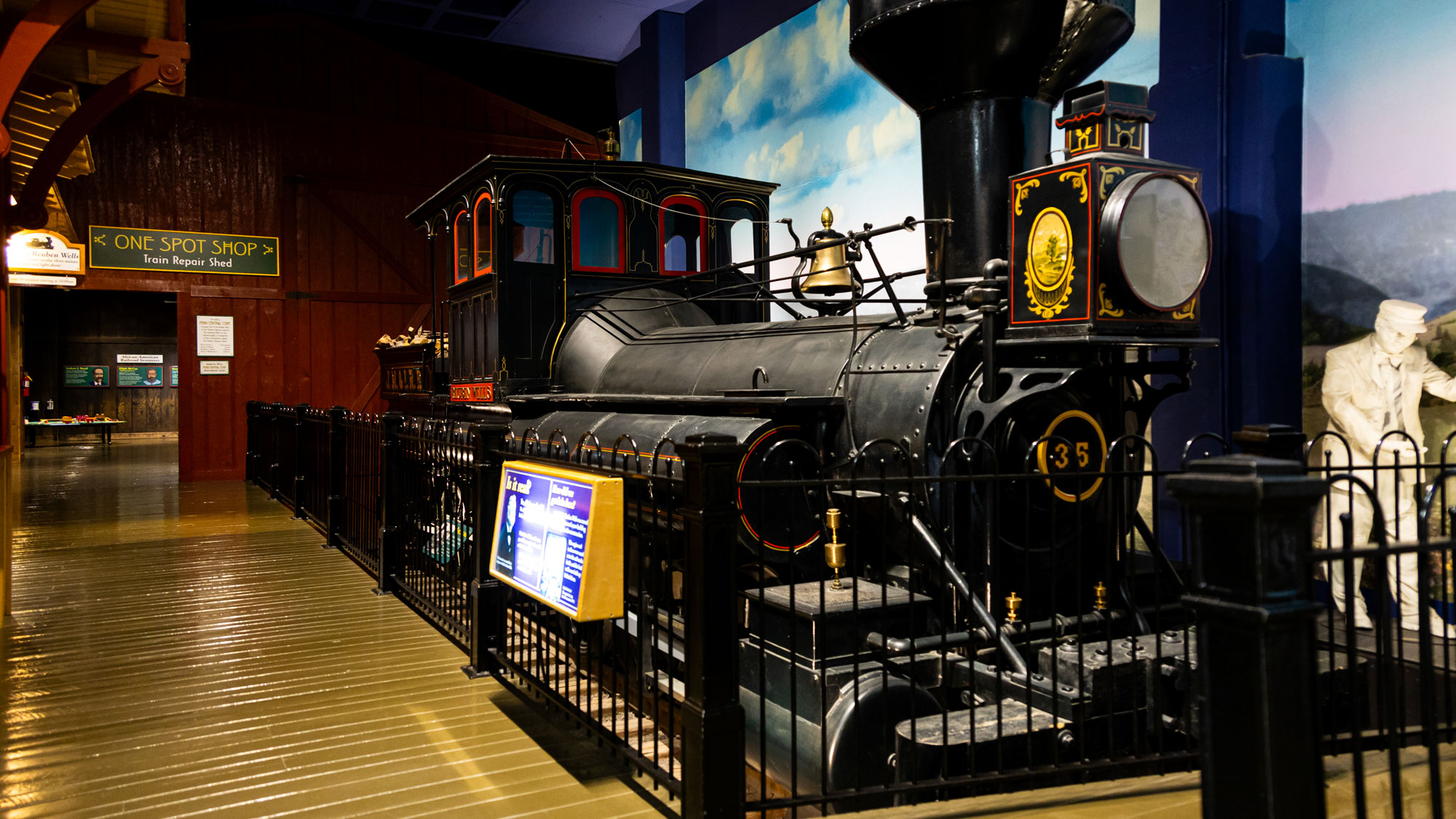 Reuben Wells locomotive with statue of conductor and entrance to train repair shop in background.