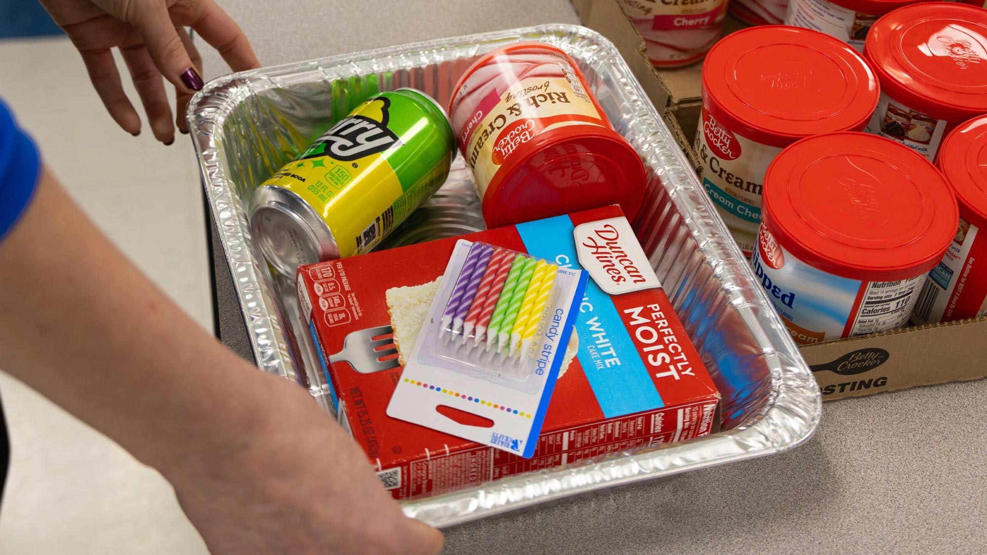 Completed cake donation kit with dry cake mix, a can of Starry, a package of candles, and a jar of frosting in an aluminum baking dish.