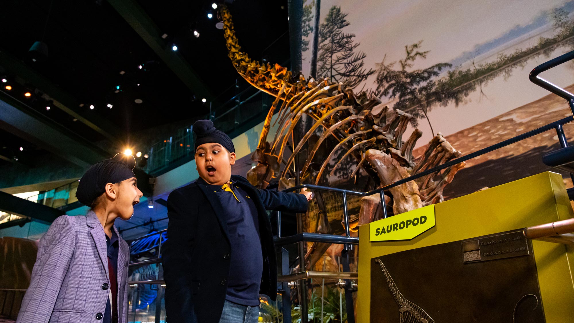Two children standing under a large sauropod fossil inside Dinosphere.
