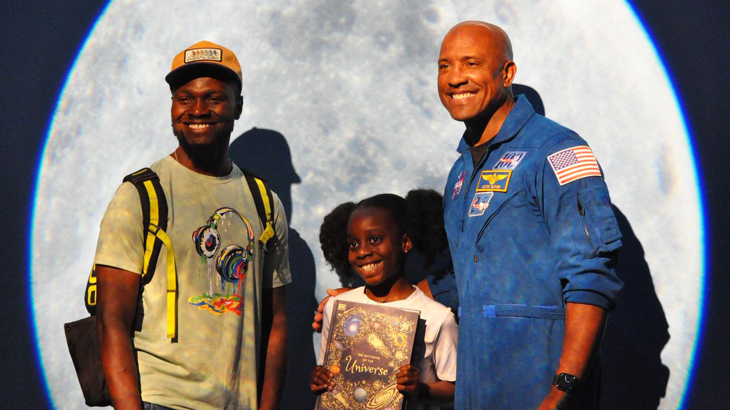 Artemis II astronaut Victor Glover posing with grown-up and child with moon in background.