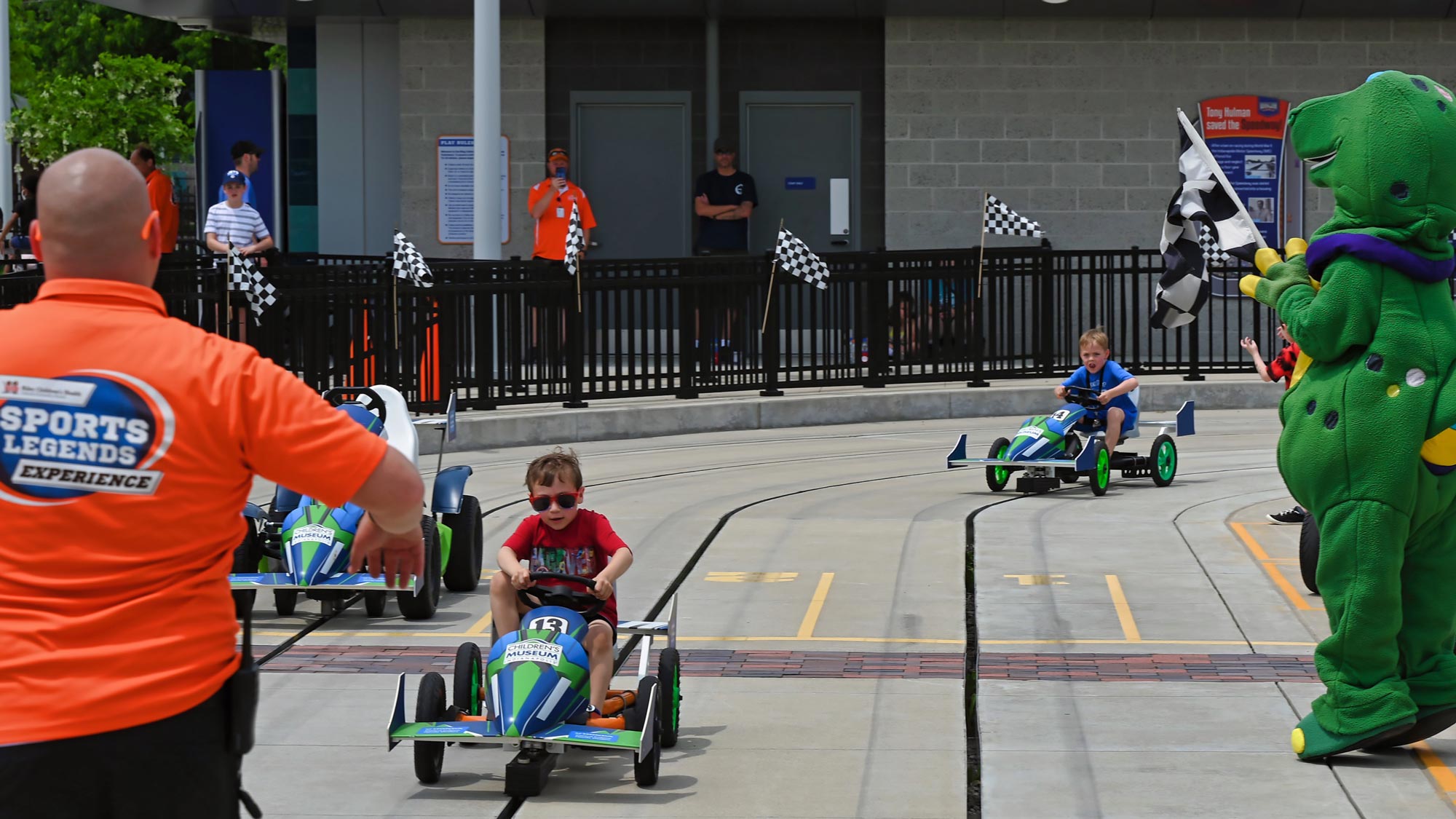 Children racing pedal cars with Rex holding a checkered flag.