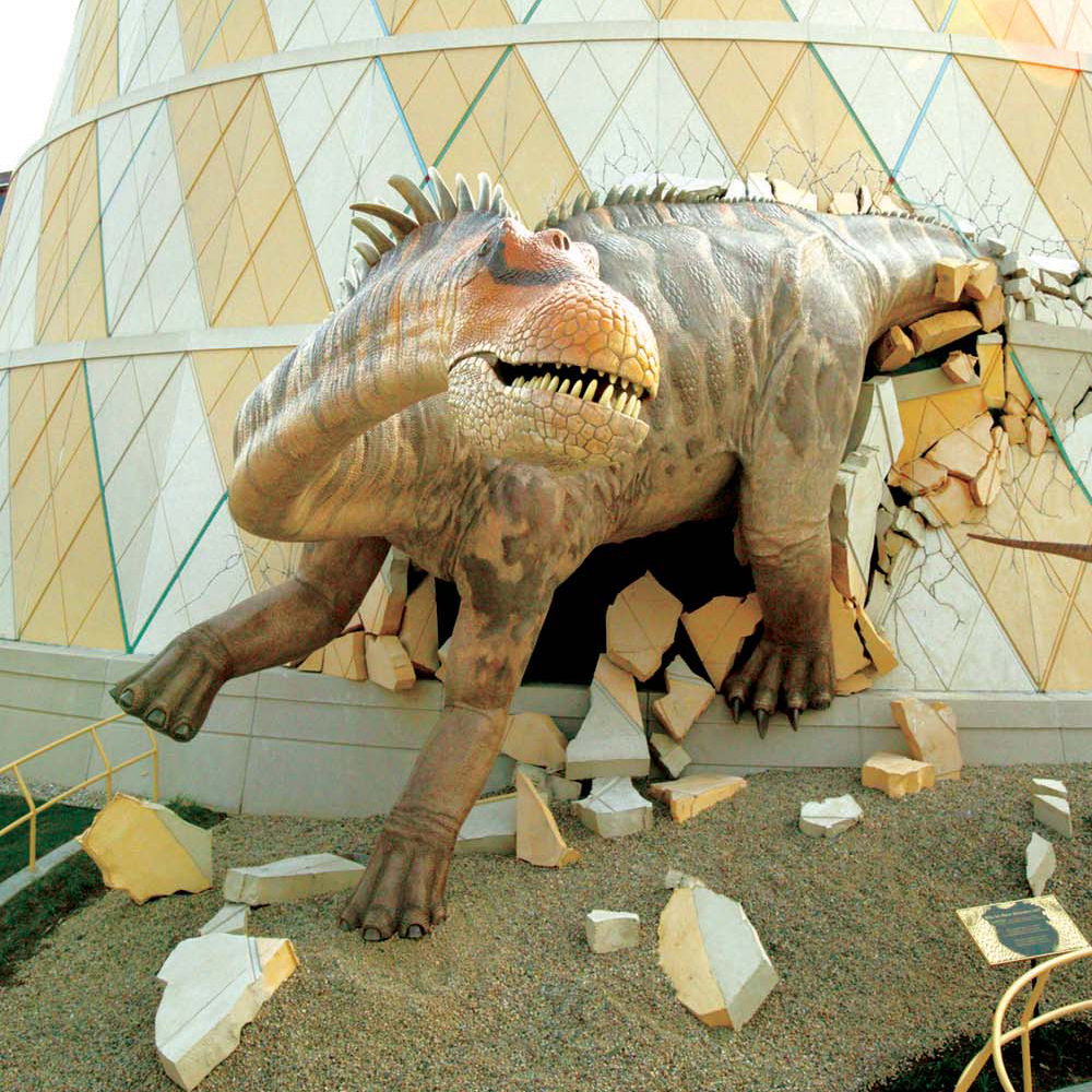 A large, realistic dinosaur sculpture breaking out through the exterior wall of The Children's Museum of Indianapolis, surrounded by shattered debris on the ground.