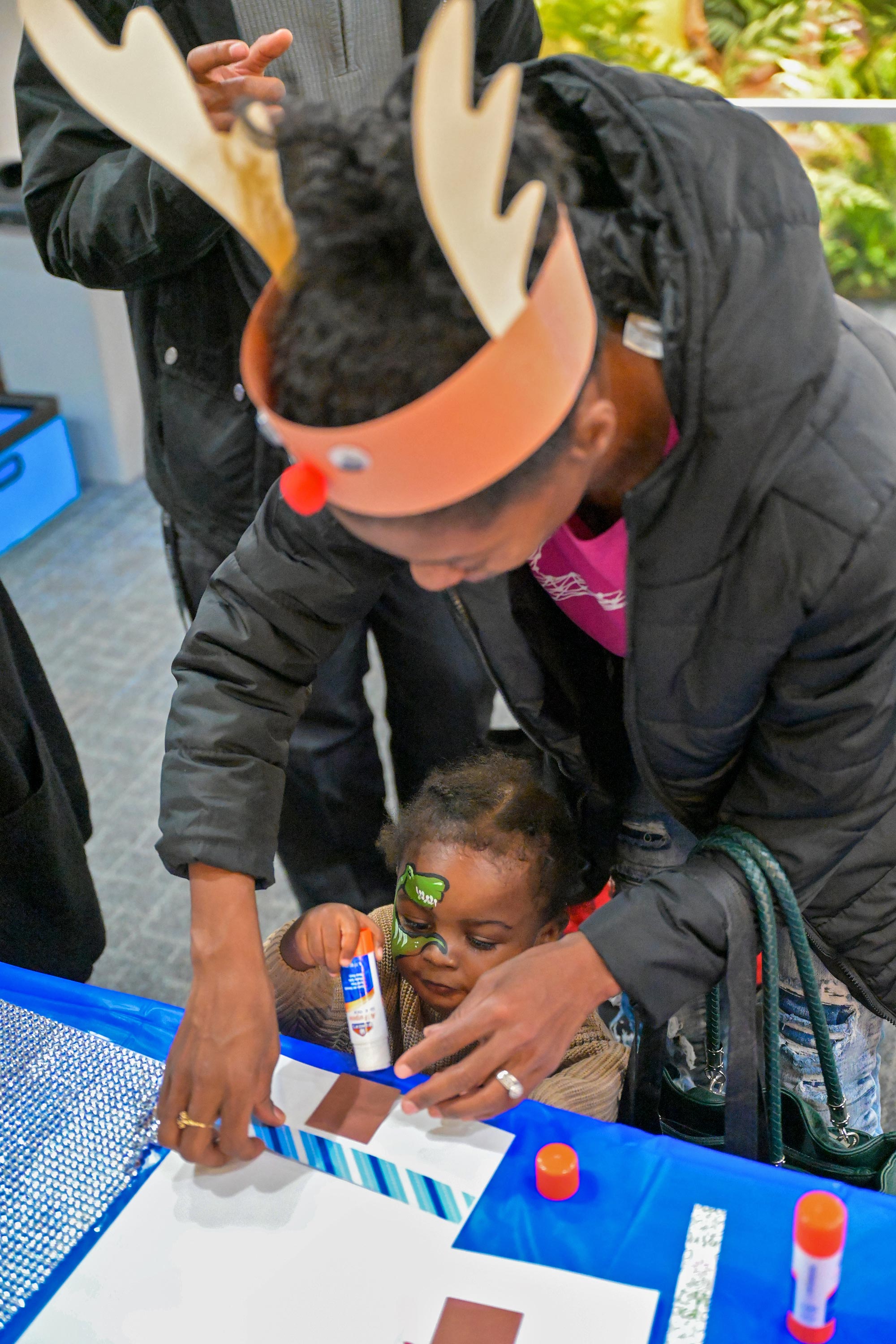 Grown-up wearing a reindeer hat and helping a child with a craft.