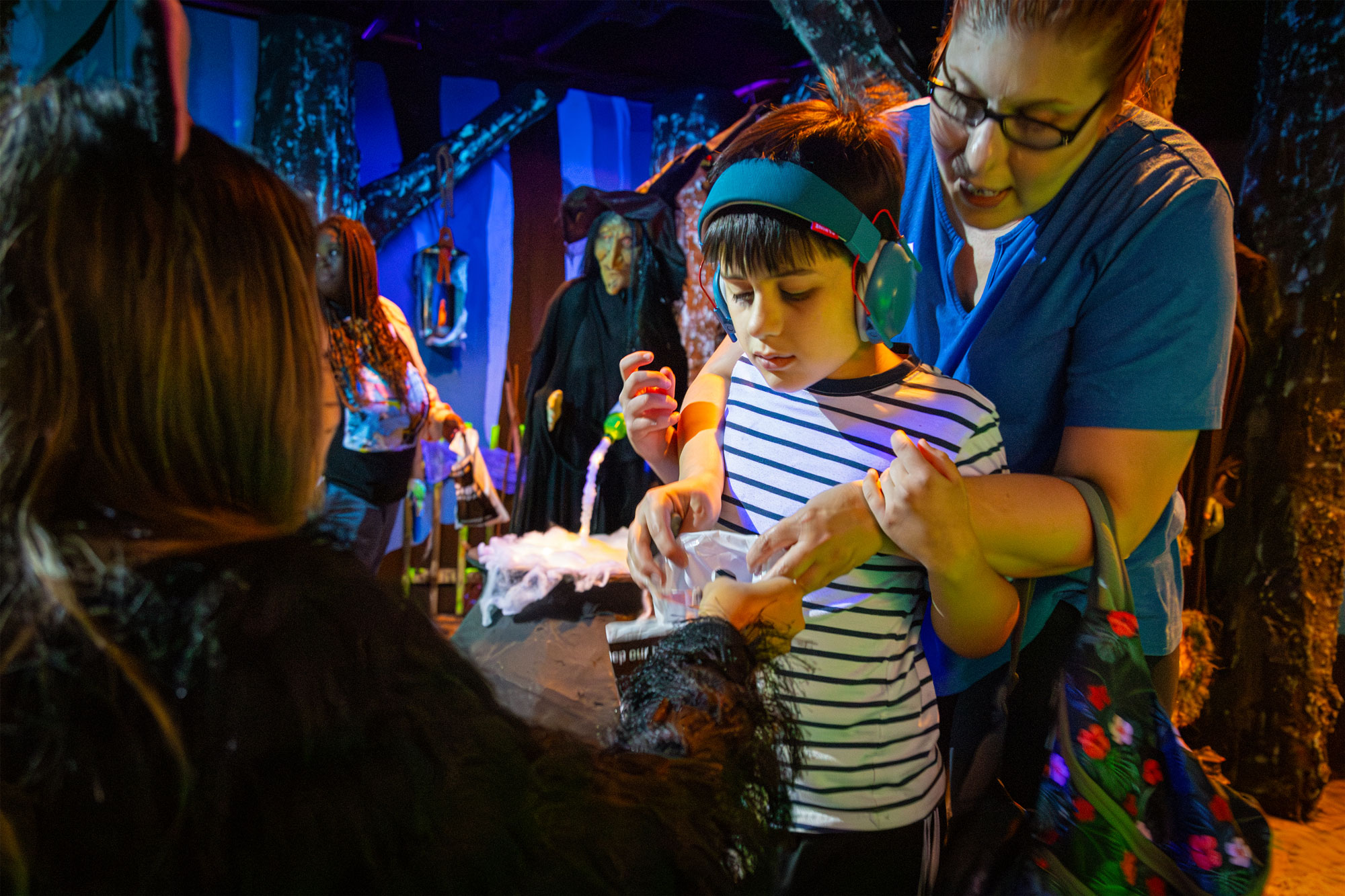 Child wearing noise canceling headphones receiving candy from a friendly witch.