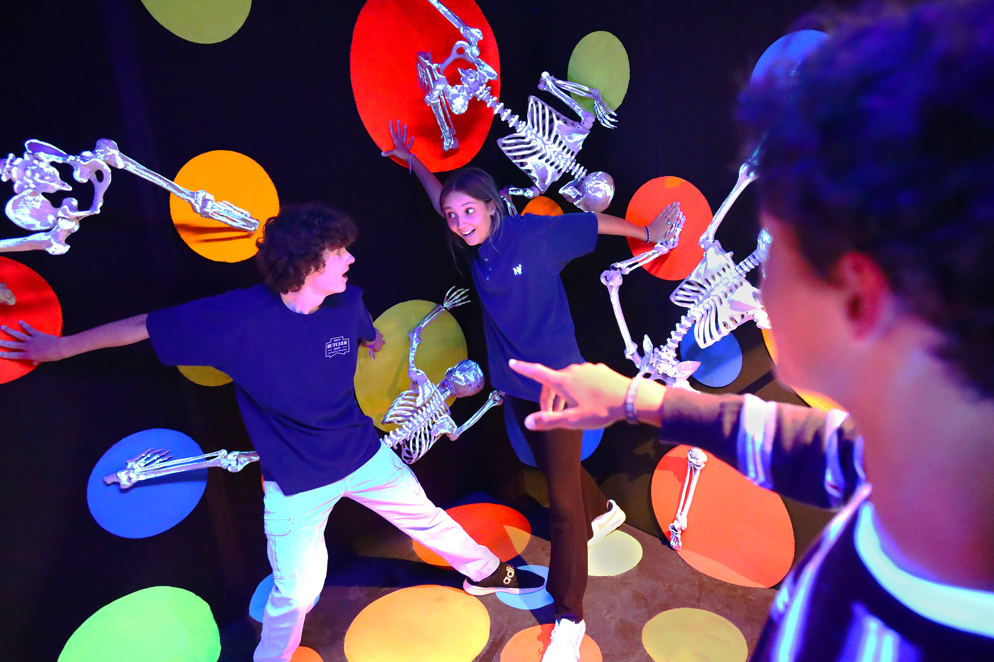 Teenagers standing in front of black wall with large colorful dots and floating skeletons.