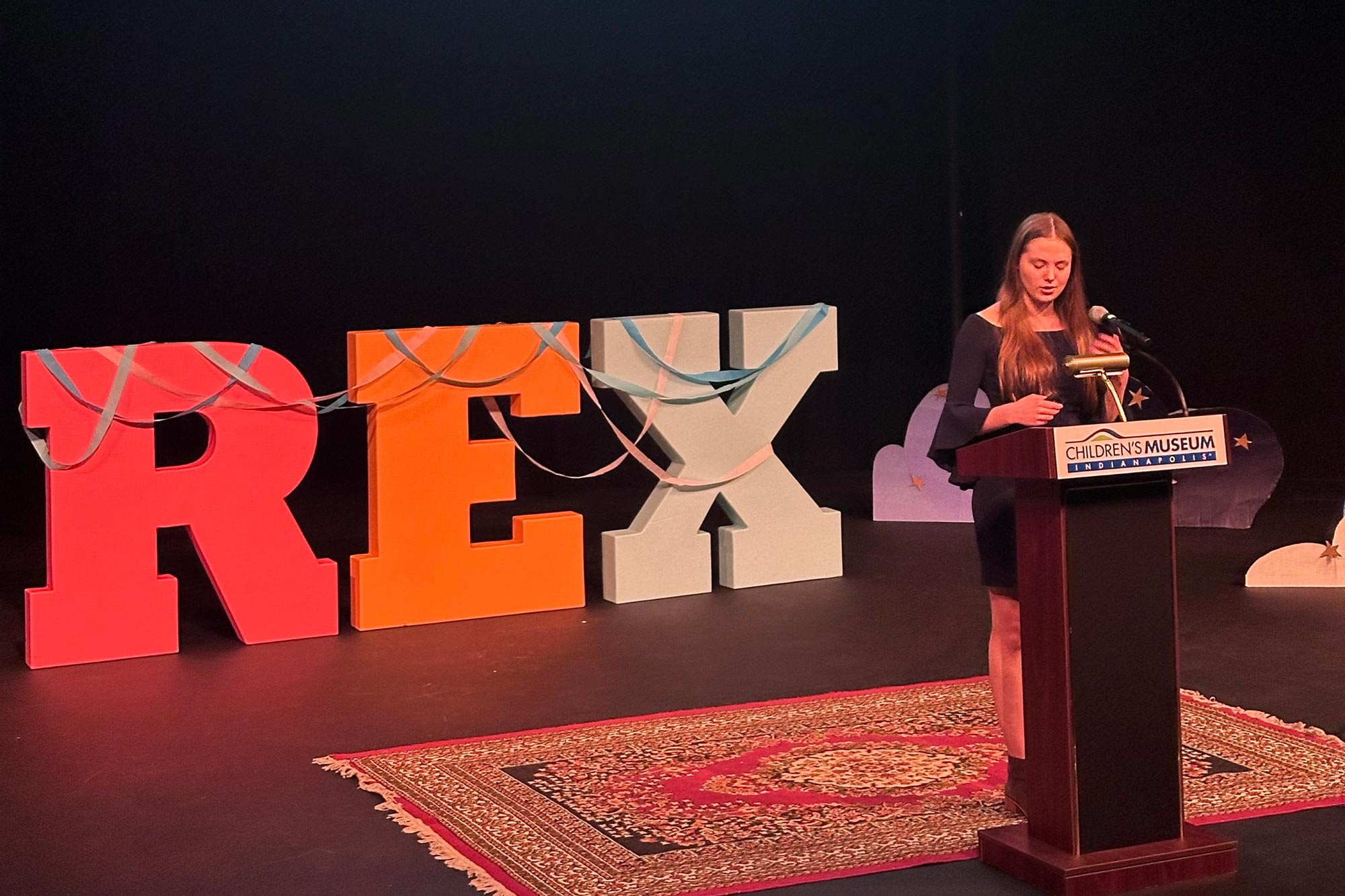 Savannah behind a podium. Giant multicolored letters behind her on stage spell Rex.