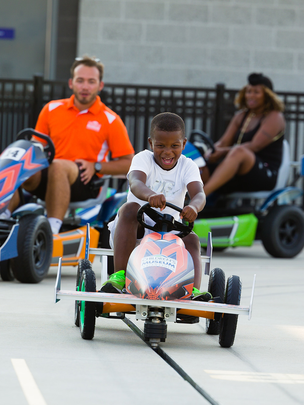 Child driving pedal car with two adults racing pedal cars in the background.