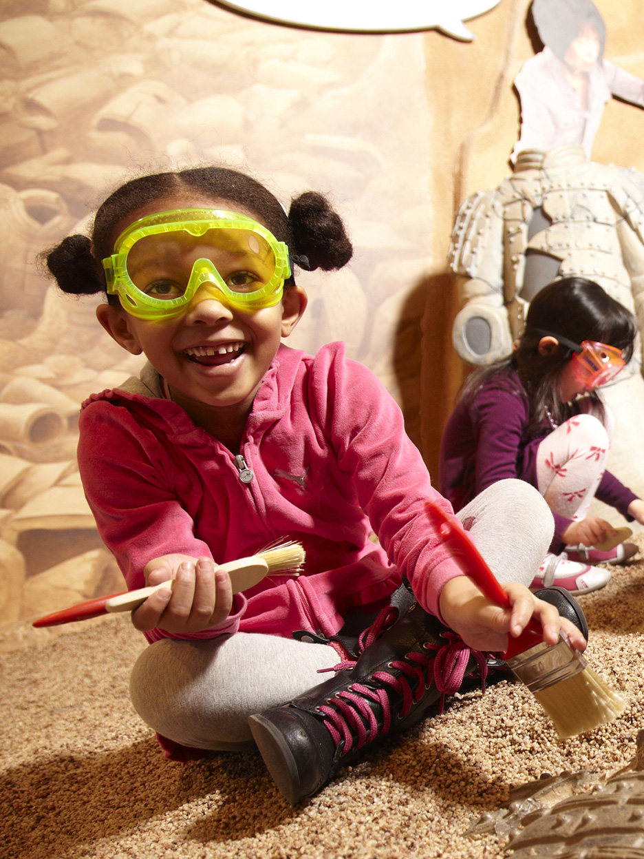 Child wearing safety goggles and digging in a pretend play dig pit.