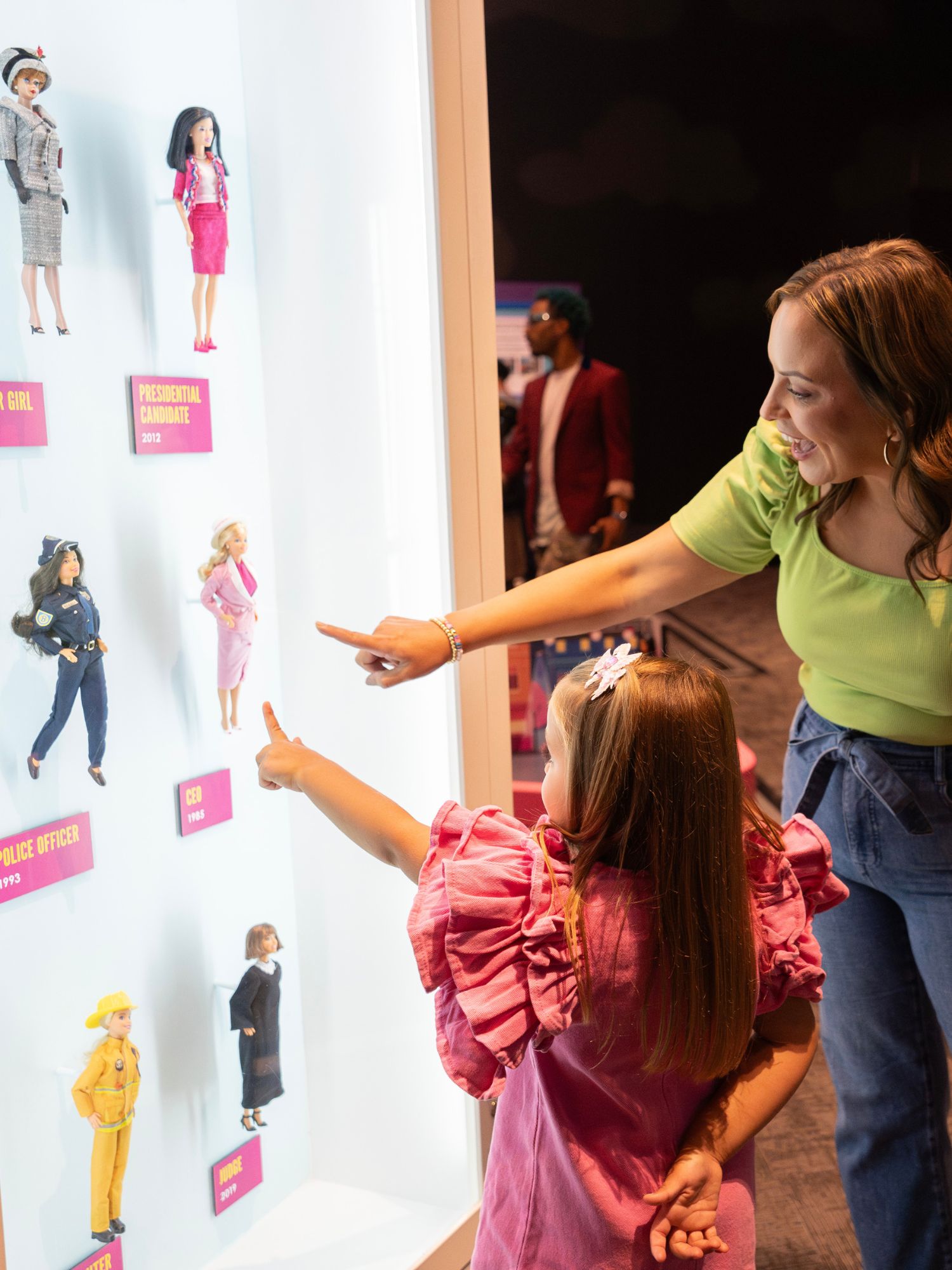 Grown up and child pointing at a display case full of Barbie dolls from history.