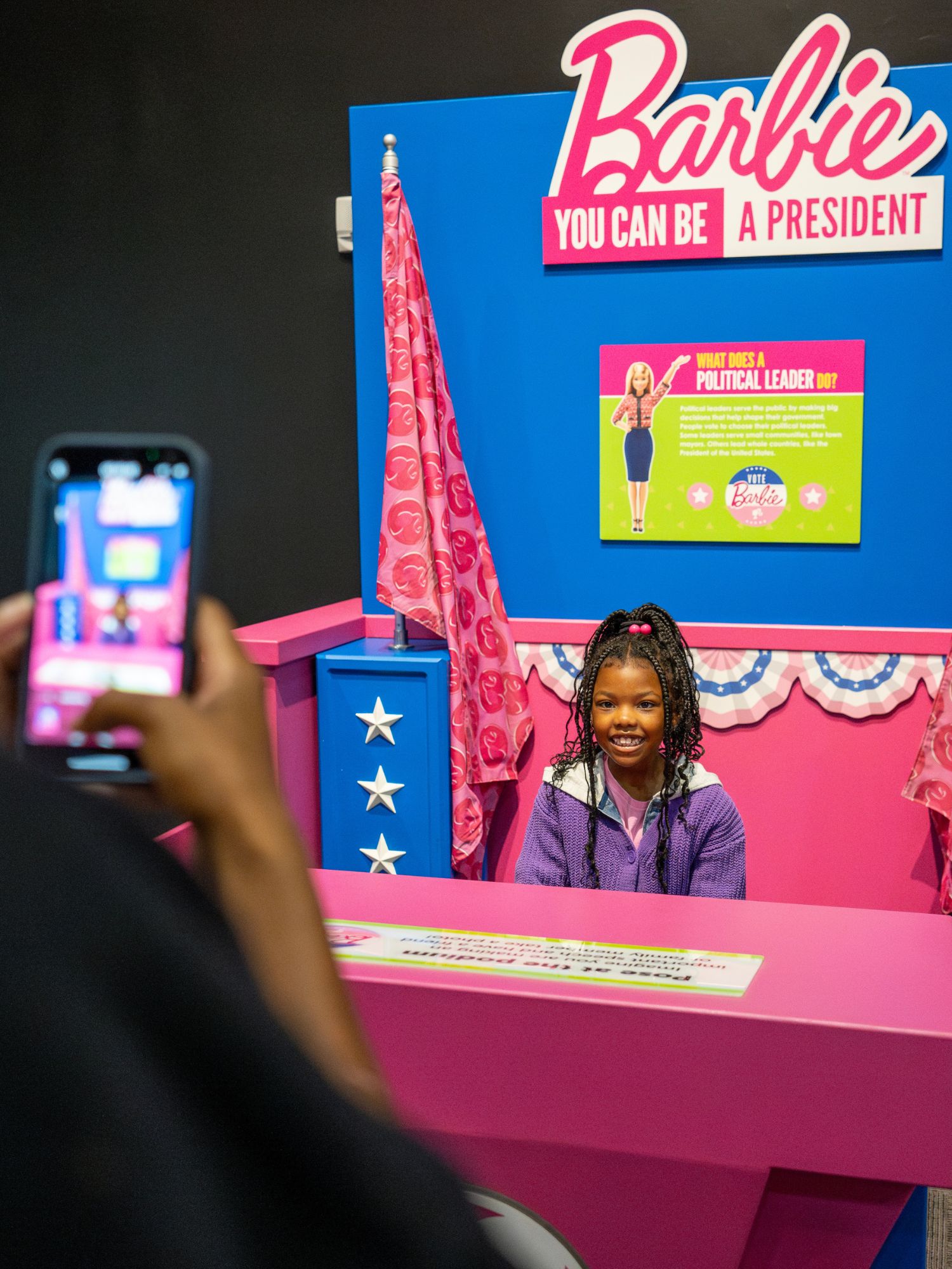 Grown-up taking a photo of child standing at a podium with a sign that says Barbie: you can be a president.