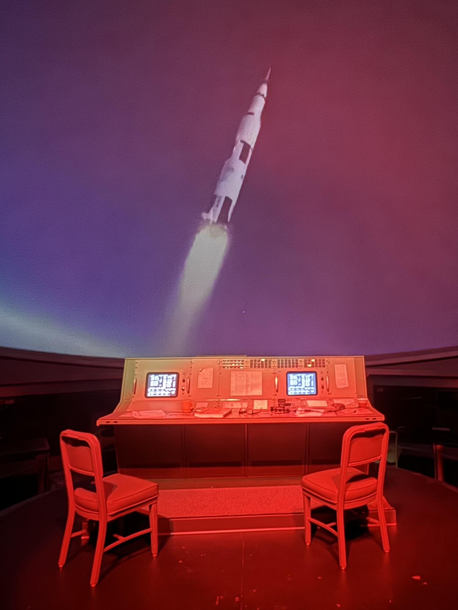 Projection of Saturn V rocket launching behind Apollo mission control console.