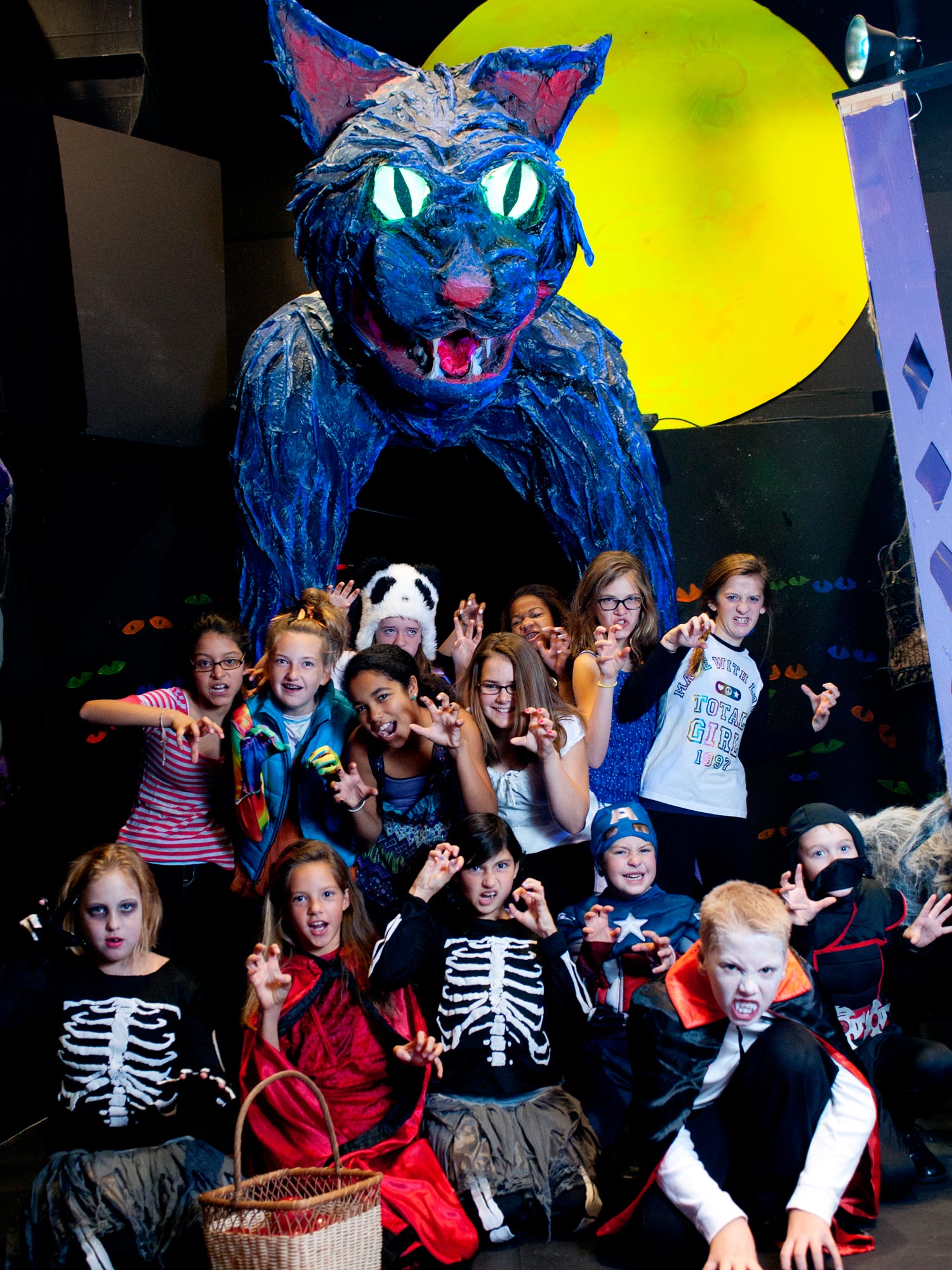 Children wearing costumes and posing in front of giant black cat and full moon.