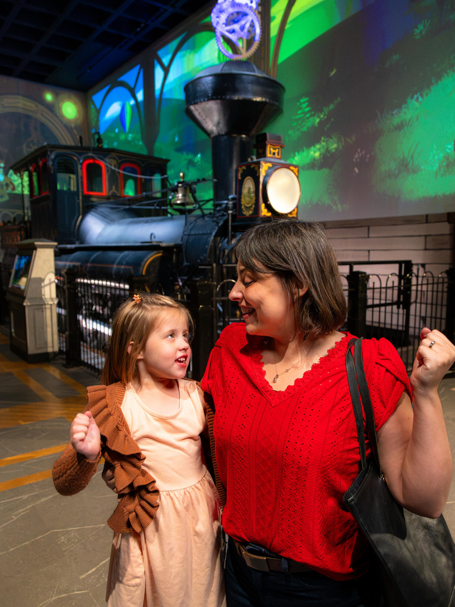 Grown-up and child flexing in front of Reuben Wells steam engine inside All Aboard! exhibit.
