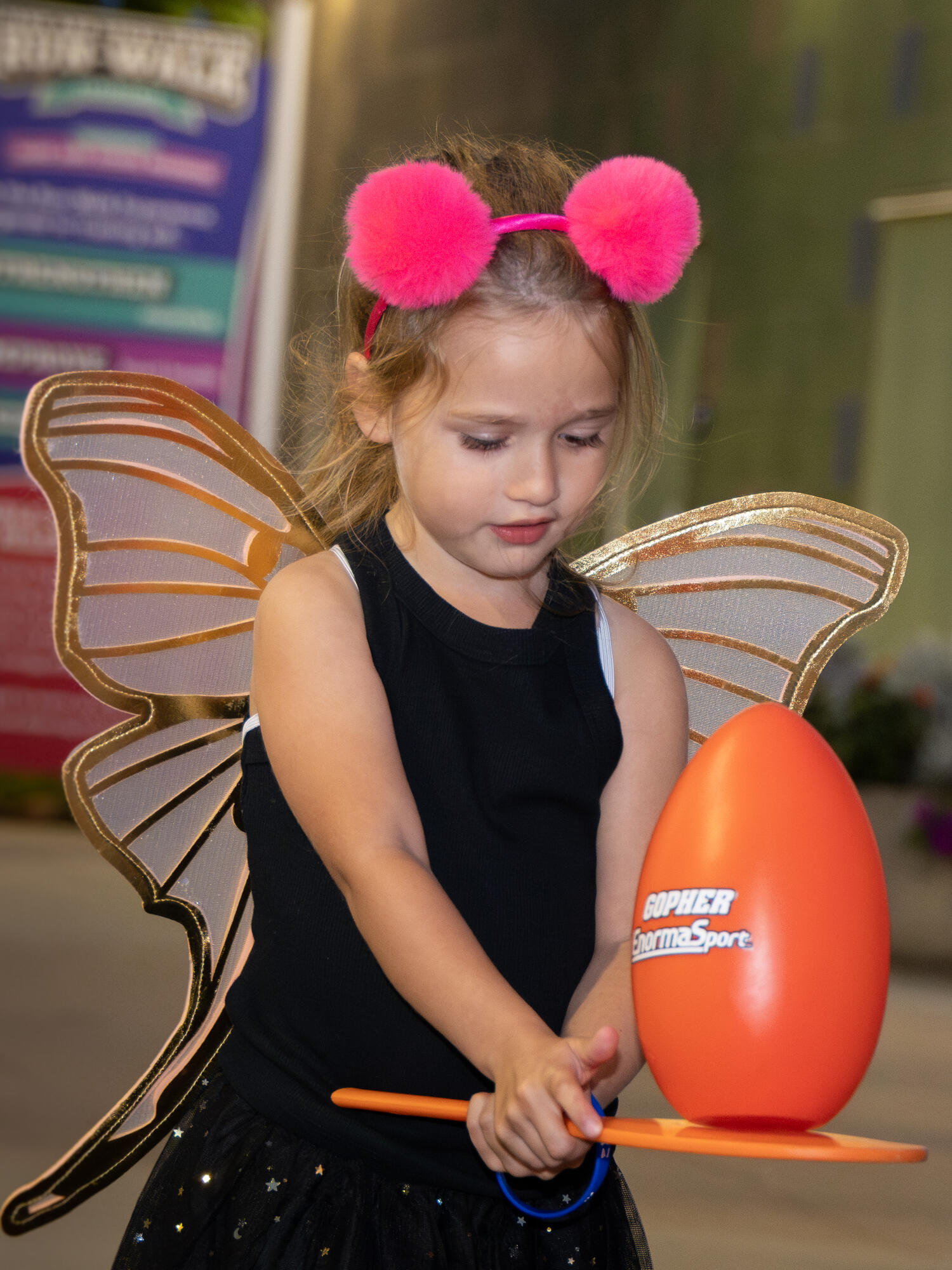 Child wearing butterfly wings and balancing a ball on a board.