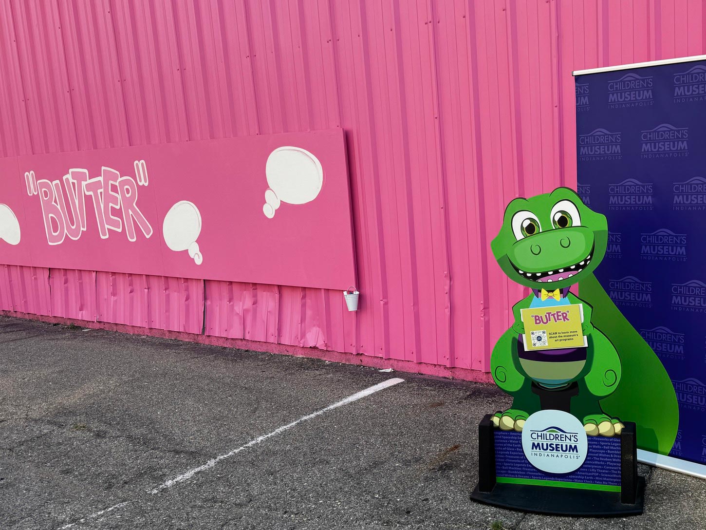 Photo opp of museum mascot Rex in front of a pink wall that has the word "BUTTER" painted on it.