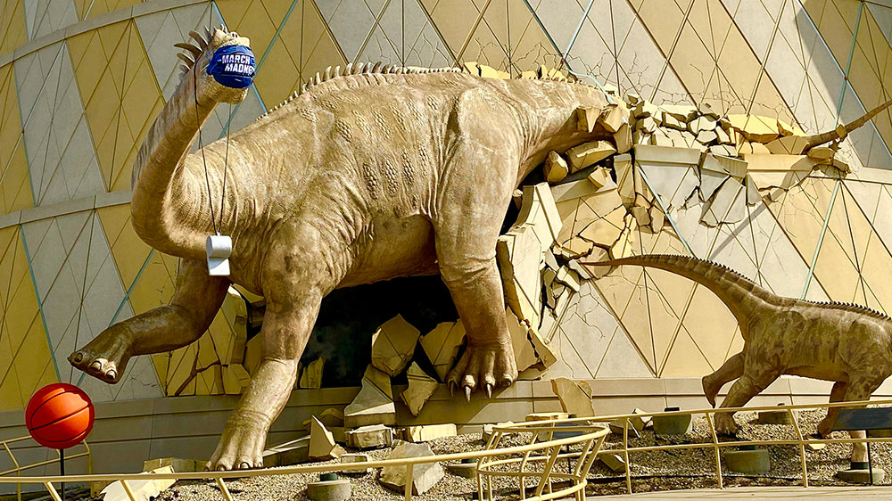 Dinos bursting out of The Children's Museum of Indianapolis with a basketball and wearing March Madness face coverings