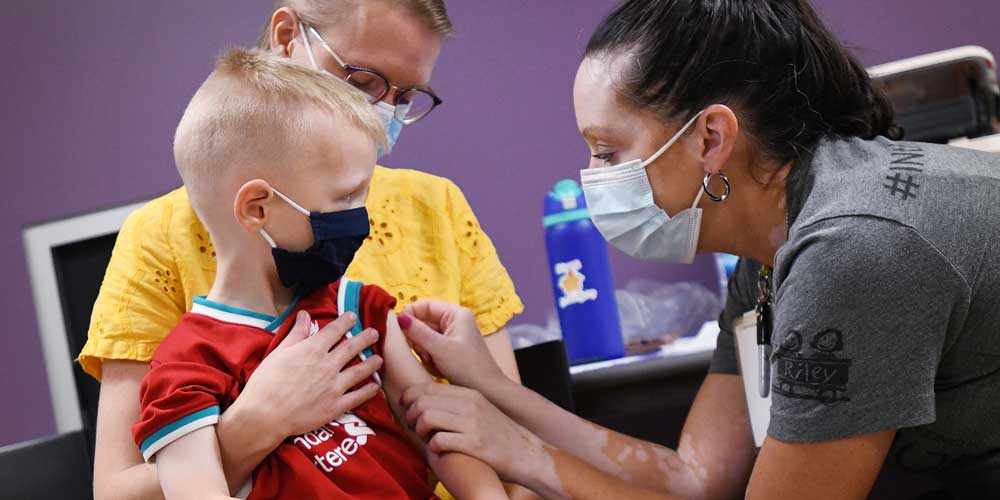 Masked child receiving a bandage on arm after receiving a shot at a Riley Children's at Indiana University Health vaccination clinic at The Children's Museum of Indianapolis.