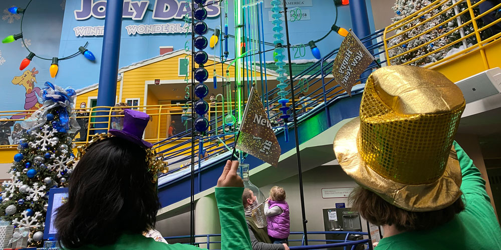 People wearing party hats and wearing Happy New Year flags in front of the Water Clock during Countdown to Noon inside the Sunburst Atrium at The Children's Museum of Indianapolis.
