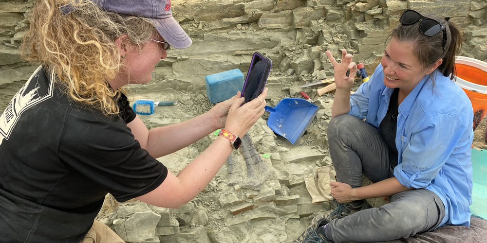 Dr. Jenn Anne uses a  phone to take a photo of Dr. Victoria Egerton holding a fossil at the Jurassic Mile dig site.
