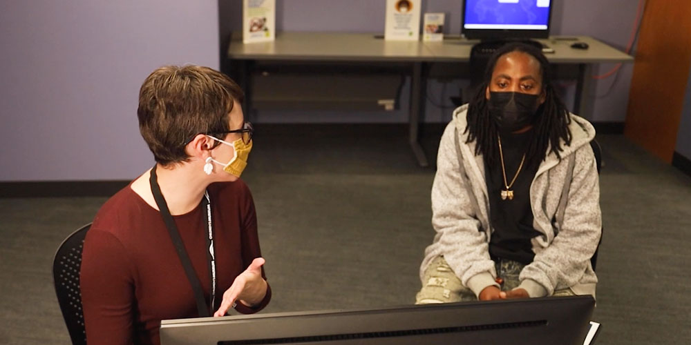 Two people wearing face masks and talking about results on a computer screen.