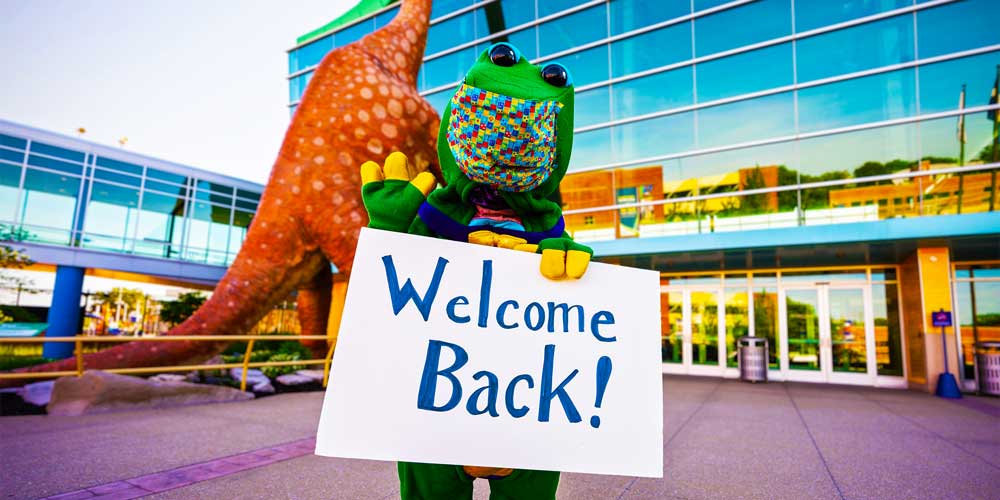 Rex wearing a mask and holding a Welcome Back sign in front of The Children's Museum of Indianapolis