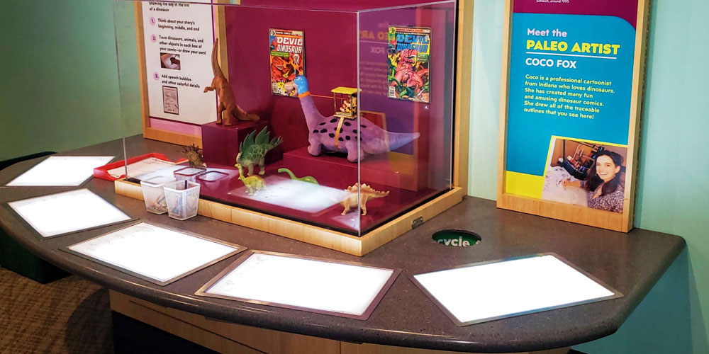 A table with backlit screens and art supplies. Comic-book style art is on display in the center and a brief bio and profile picture of Coco Fox is on the right.