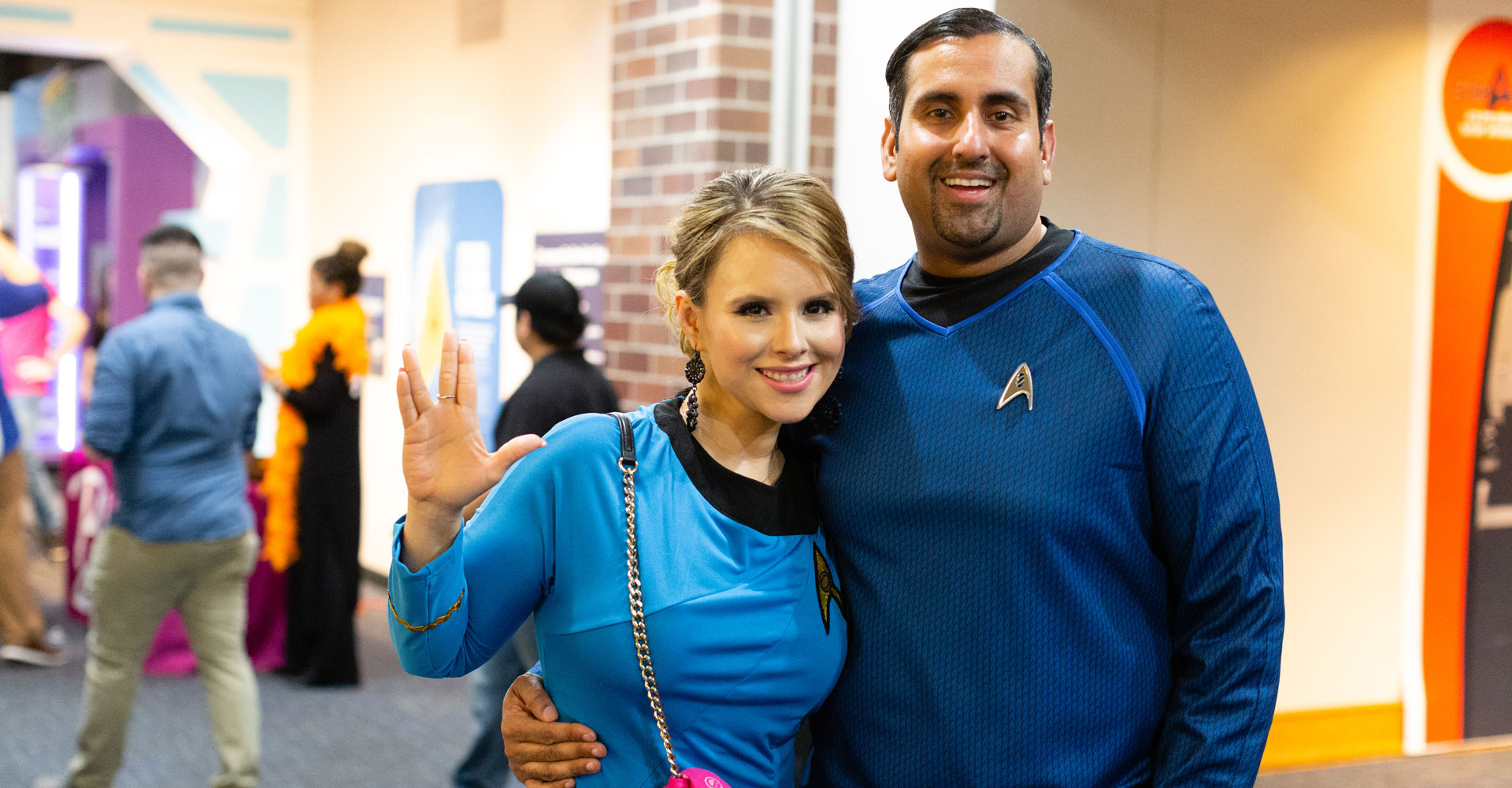 Man and woman wearing blue Star Trek costumes. Woman is making the Vulcan salute with her hand.