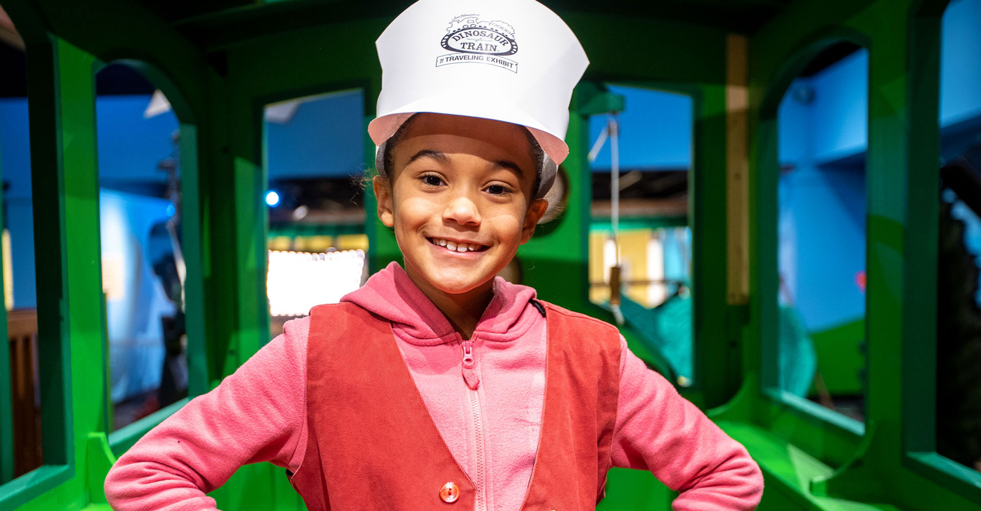 Child wearing pink and wearing a white Dinosaur Train conductor cap standing inside the cab of a replica of the Dinosaur Train.