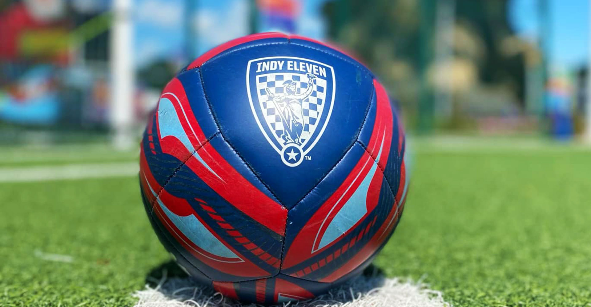 Red, blue, white Indy Eleven soccer ball sitting on a green soccer pitch.