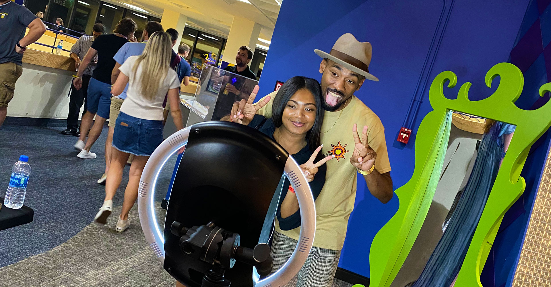 Woman wearing Silent Disco headphones and a man wearing a baseball cap, smiling and posing for a selfie in front of a T. rex inside Dinosphere.