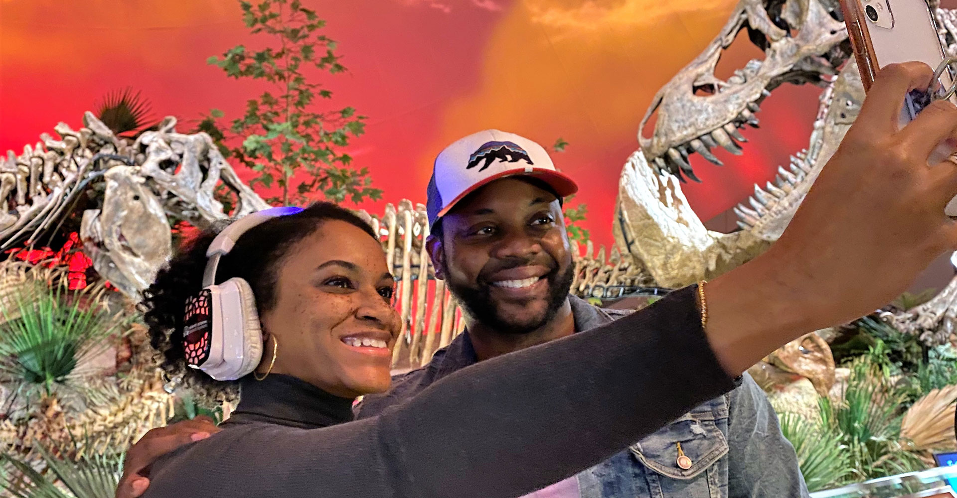 Woman wearing Silent Disco headphones and a man wearing a baseball cap, smiling and posing for a selfie in front of a T. rex inside Dinosphere.