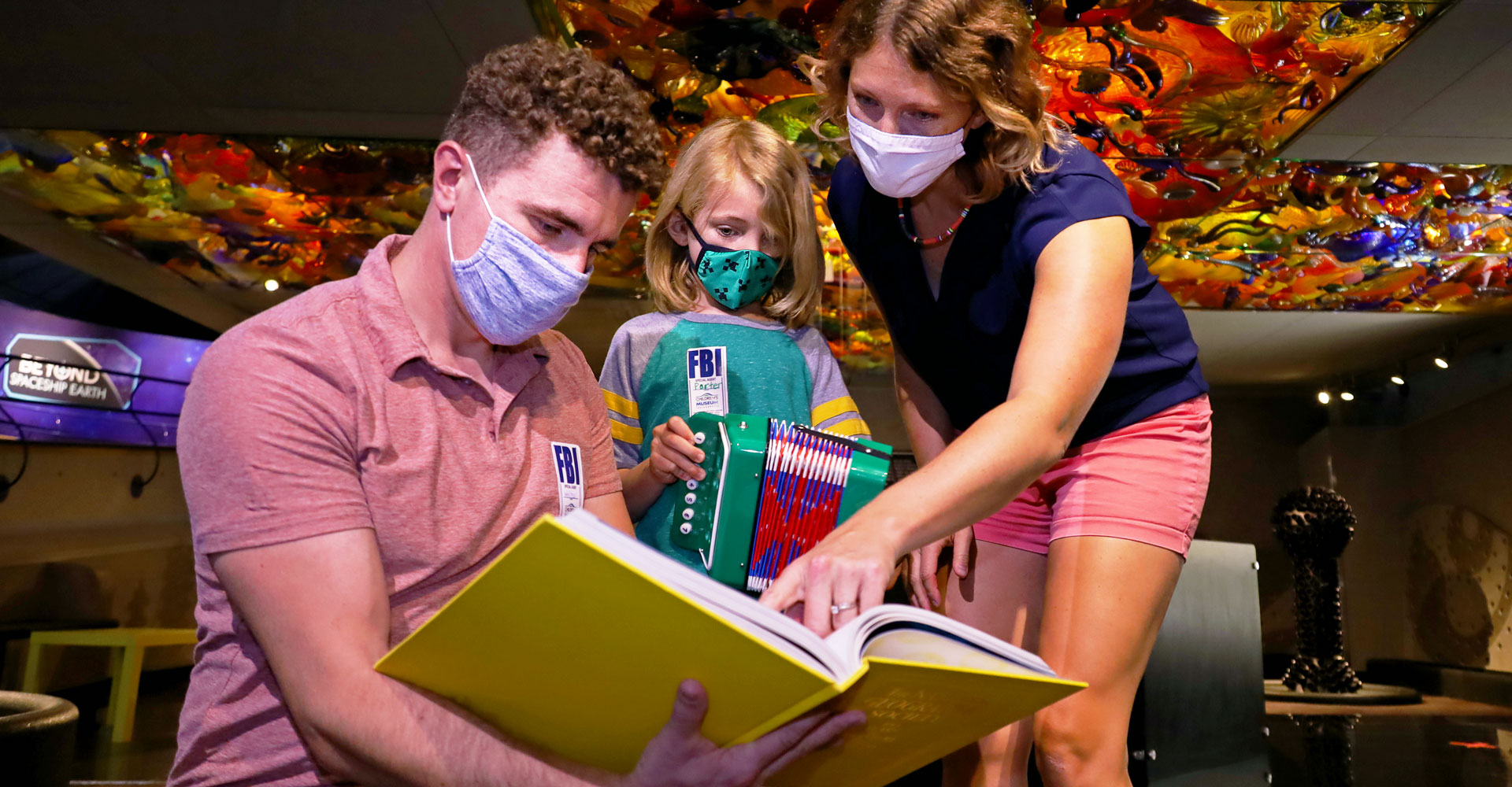 Two adults and a child wearing masks and looking in a book in the Fireworks of Glass exhibit