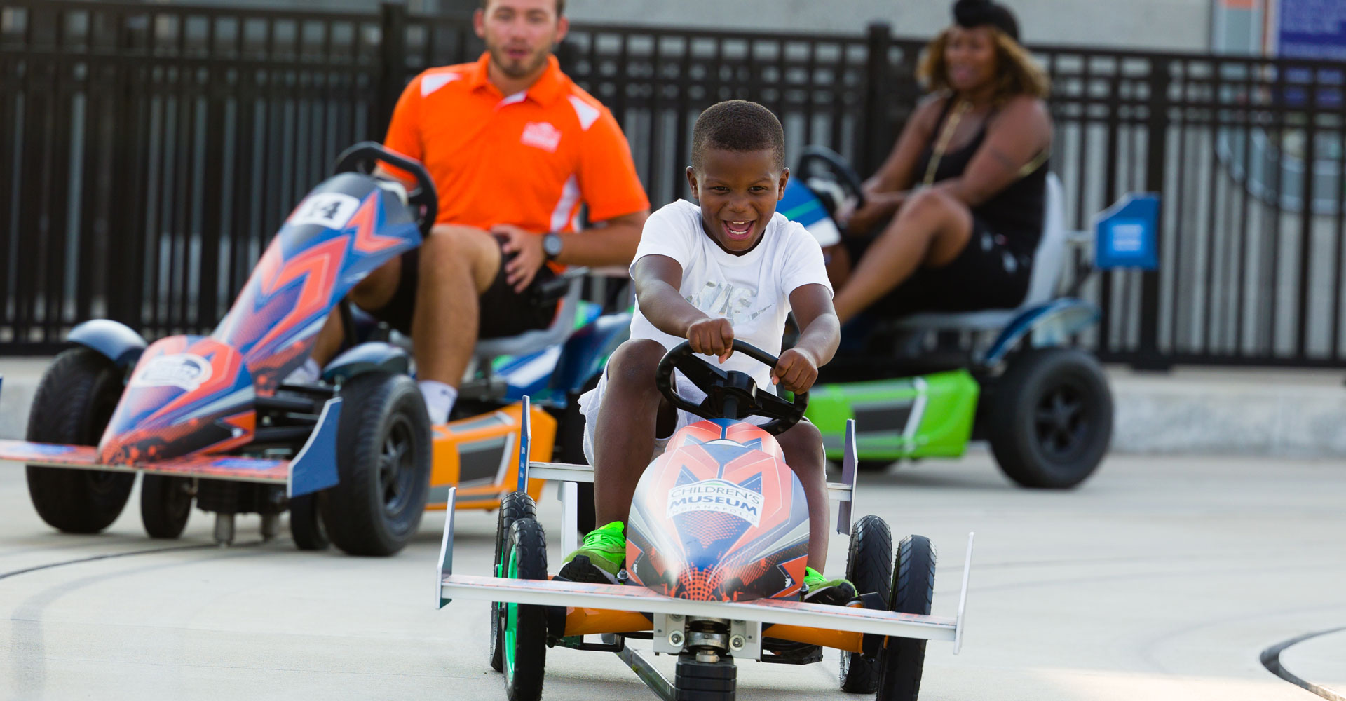 Child driving pedal car with two adults racing pedal cars in the background.