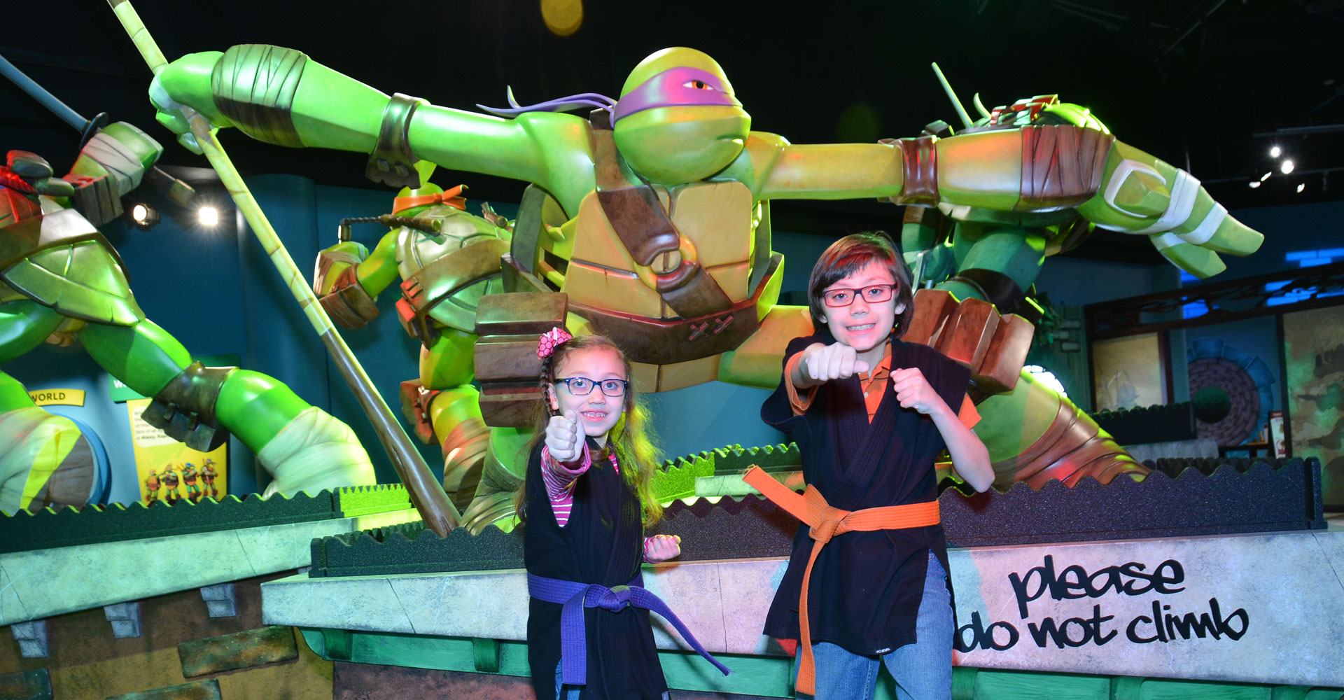 Two older children posing in front of large sculptures of the Teenage Mutant Ninja Turtles.
