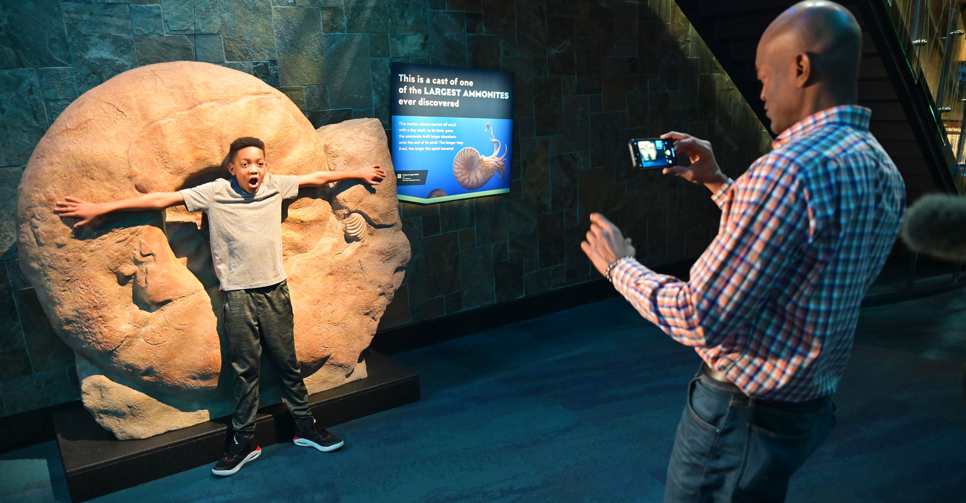 Grown-up taking a photo of child with mouth open and arms spread out, showing the size of a giant ammonite fossil.