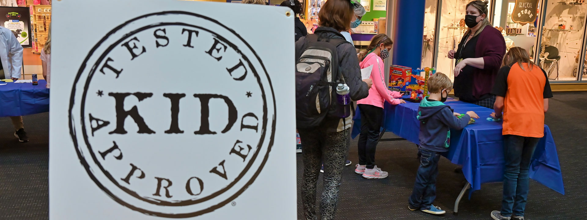 Kid-Tested, Kid-Approved testing sign and families playing with toys outside The Children's Museum Store.