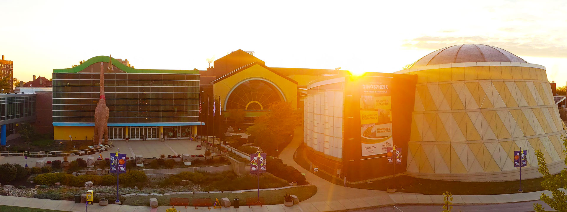 A wide shot of the outside front of the museum with the sun rising behind it. You can see the dinosaurs peeking in and the Dinosphere dome.