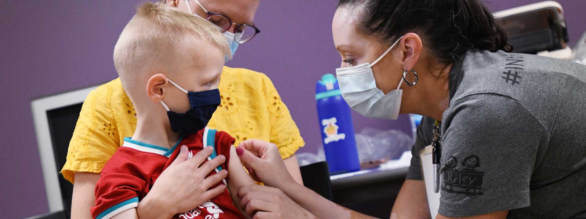 Masked child receiving a bandage on arm after receiving a shot at a Riley Children's at Indiana University Health vaccination clinic at The Children's Museum of Indianapolis.
