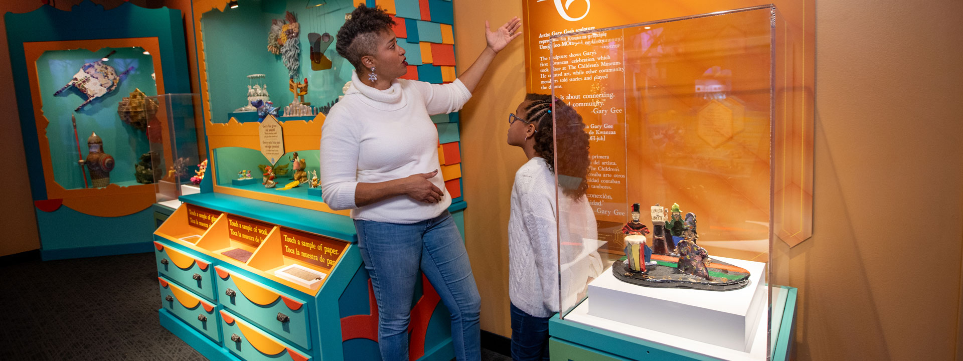 An African American mother and daughter stand in front of a label in Gerty's workshop actively engaged in reading and discussing the content which is about Kwanzaa.