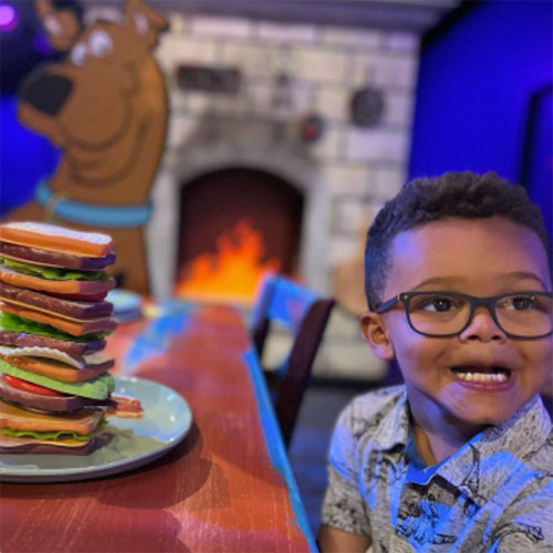A child is sitting near a table and looking off-camera to the right. Pretend food is stacked on the table like a sandwich. A cutout of SCOOBY-DOO is in the background at Access Pass cultural attraction The Children's Museum of Indianapolis