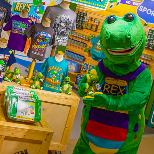 Mascot Rex standing in front of a T-shirt display in The Museum Store.